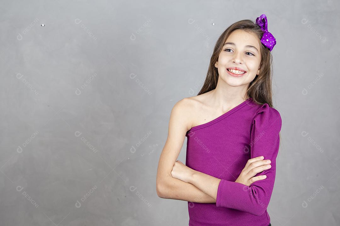 Jovem de aproximadamente dez anos com os braços cruzados sorrindo olhando para a câmera