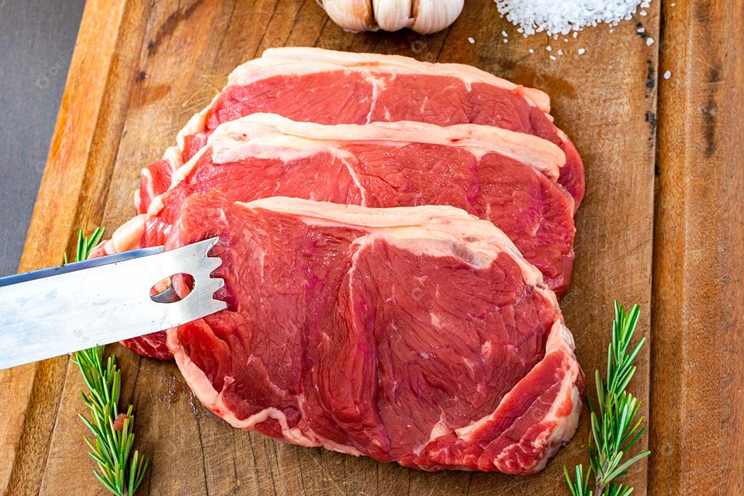 tong pegando carne vermelha crua, bife do lombo com gordura em uma tábua de corte junto com tomilho, alho, cebola e sal