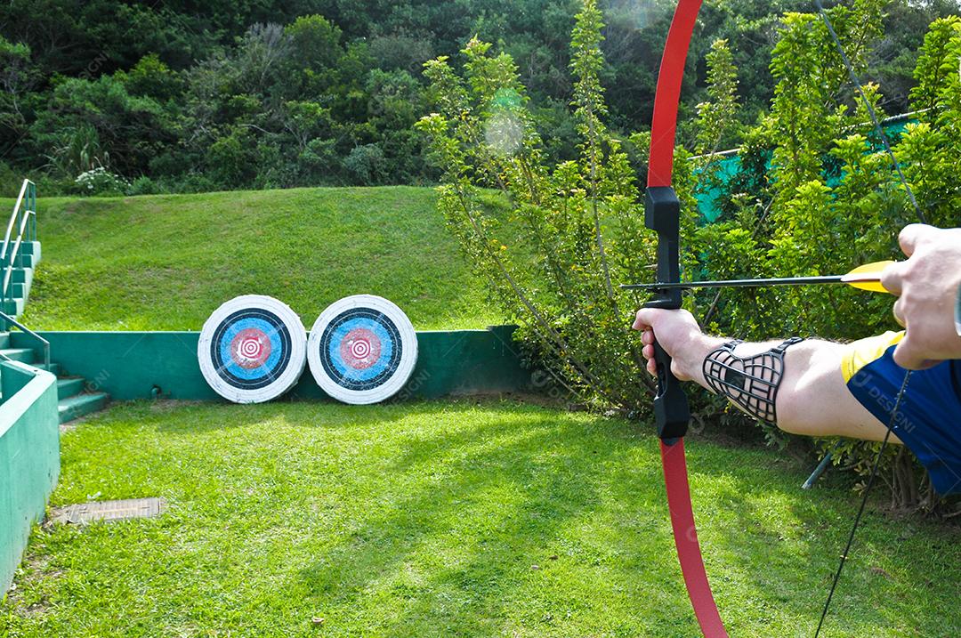 Homem atirando no alvo com arco e flecha usando todos os equipamentos de segurança deste esporte.