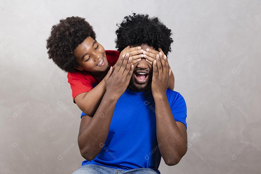 filho afro cobrindo os olhos do pai com a mão para fazer uma surpresa. Dia dos Pais