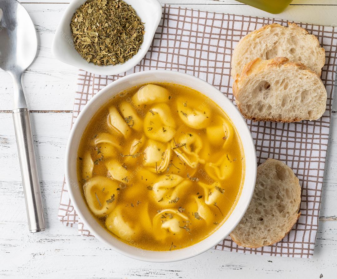 Sopa de cappelletti ou tortellini brodo em uma tigela com fatias de pão e temperos sobre mesa de madeira.