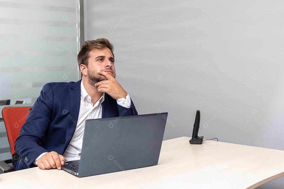 Homem de negócios jovem pensativo no escritório sentado na mesa de trabalho na frente do laptop.