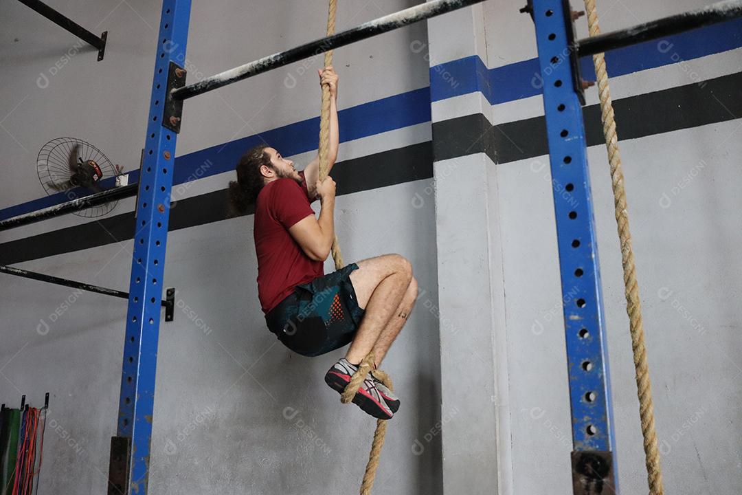 Homem praticando crossfit exercios subindo em corda sobre uma academia Conceito saúde melhor
