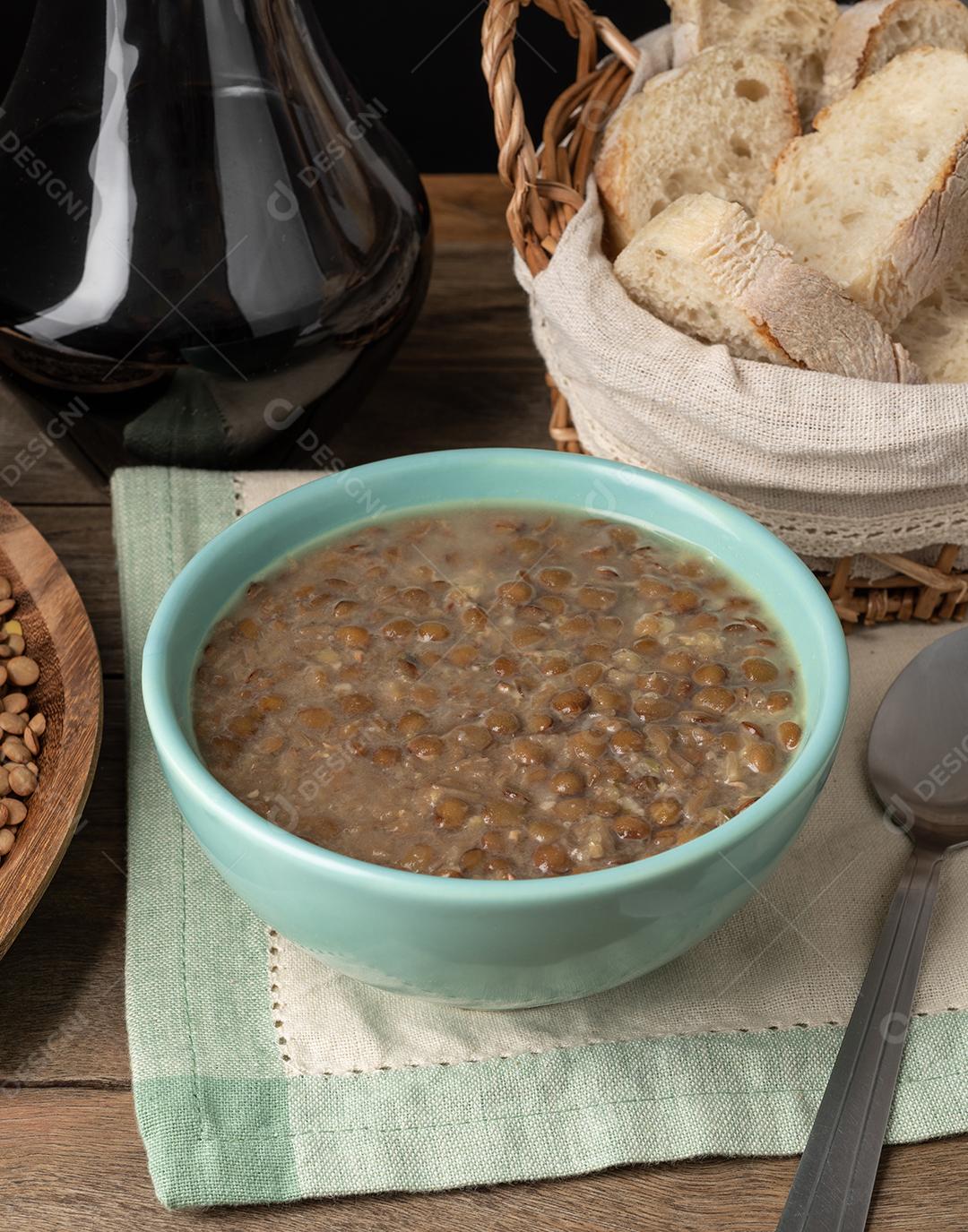Sopa de lentilha marrom em uma tigela com fatias de pão e vinho sobre a mesa de madeira.