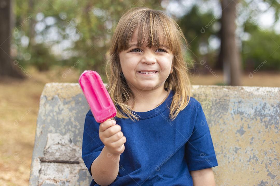 Menina caucasiana bonitinha chupando um picolé no banco do parque ao ar livre