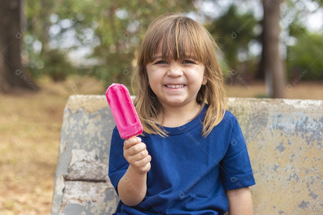 Menina caucasiana bonitinha chupando um picolé no banco do parque ao ar livre