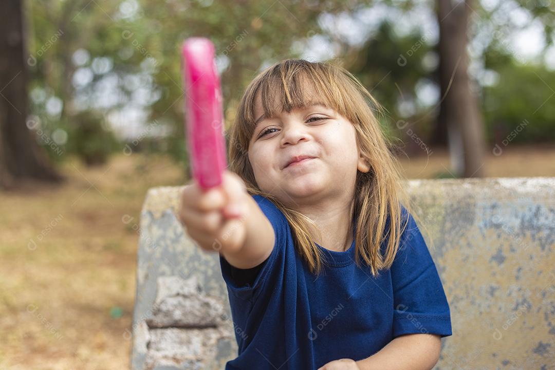 Menina caucasiana bonitinha chupando um picolé no banco do parque ao ar livre