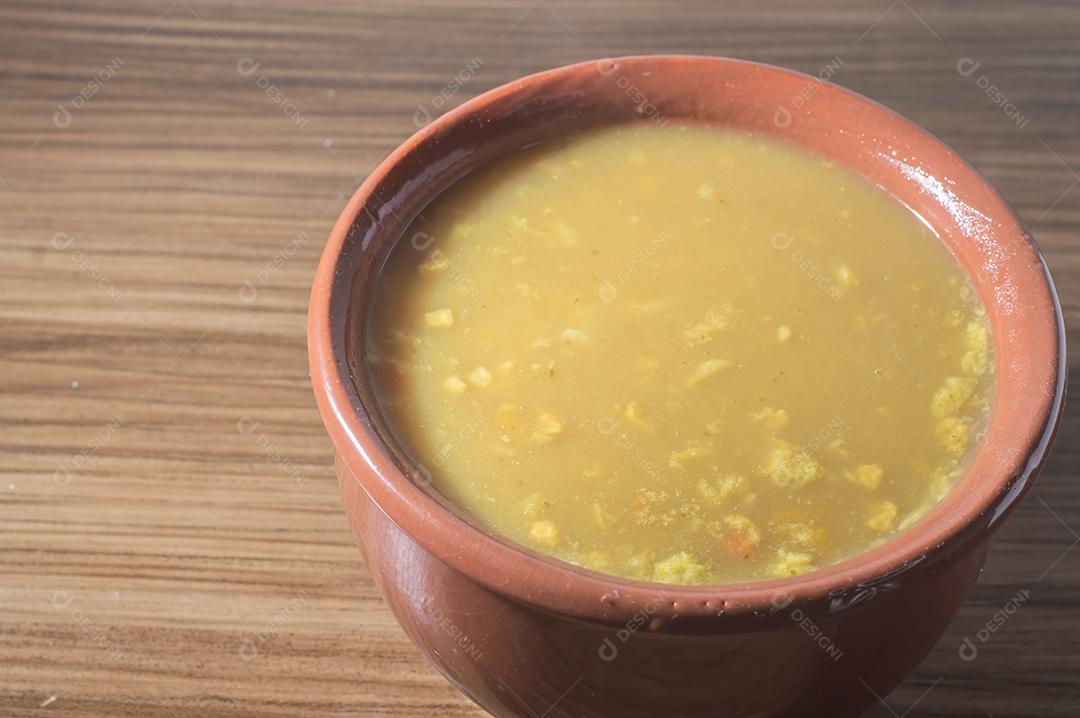 Sopa amarela em uma tigela de barro, a sopa está em uma mesa de madeira com espaço de cópia e colher.