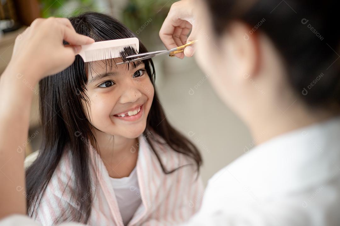 Mãe asiática cortando cabelo para sua filha na sala de estar em casa