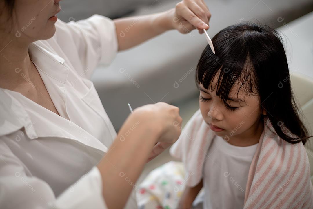 Mãe asiática cortando cabelo para sua filha na sala de estar em casa