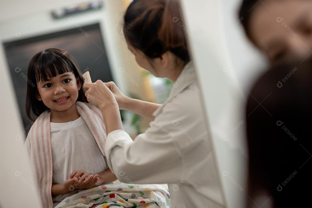 Mãe asiática cortando cabelo para sua filha na sala de estar em casa