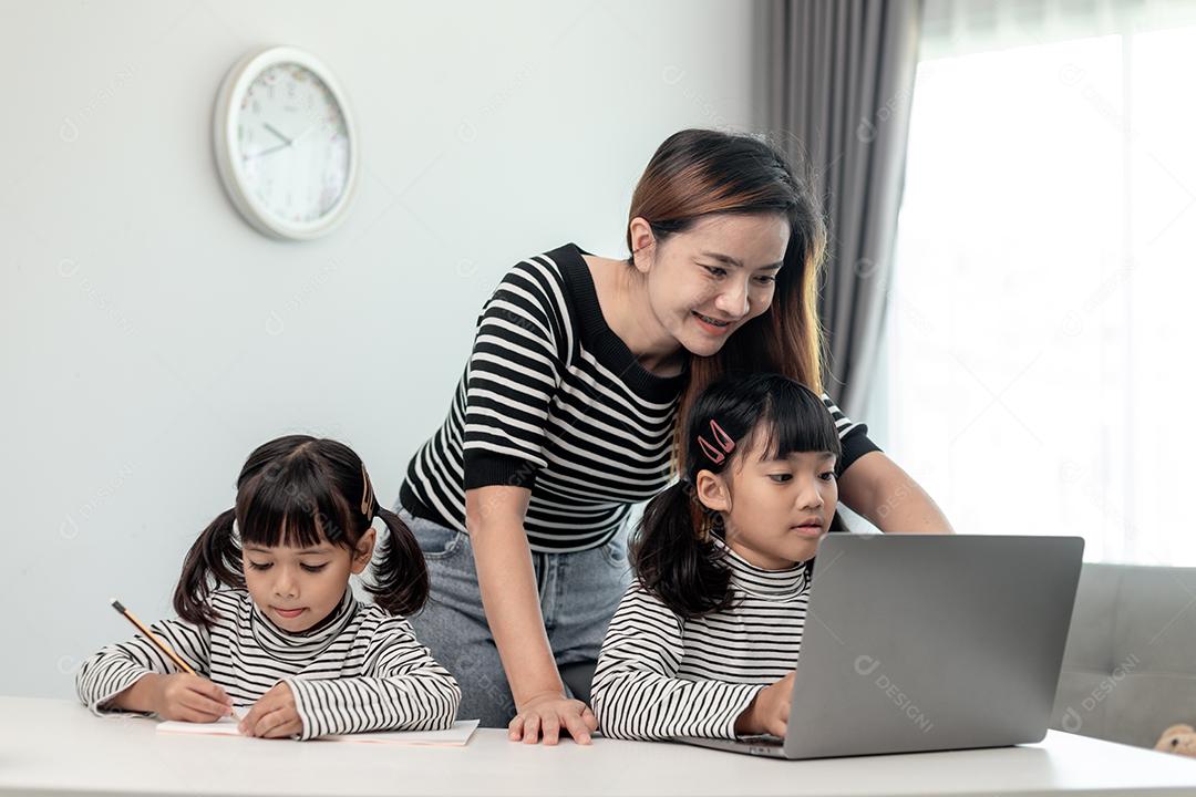 Garotinha asiática aprendendo aula online em casa com a mãe. A criança pré-escolar usa o laptop para fazer a lição de casa, o ensino em casa do professor da escola pela internet remota digital com o apoio da mãe.