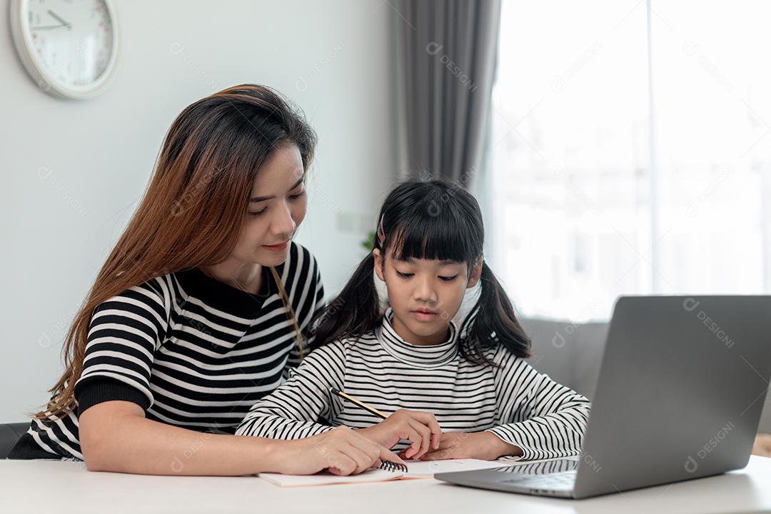 Garotinha asiática aprendendo aula online em casa com a mãe. A criança pré-escolar usa o laptop para fazer a lição de casa, o ensino em casa do professor da escola pela internet remota digital com o apoio da mãe.