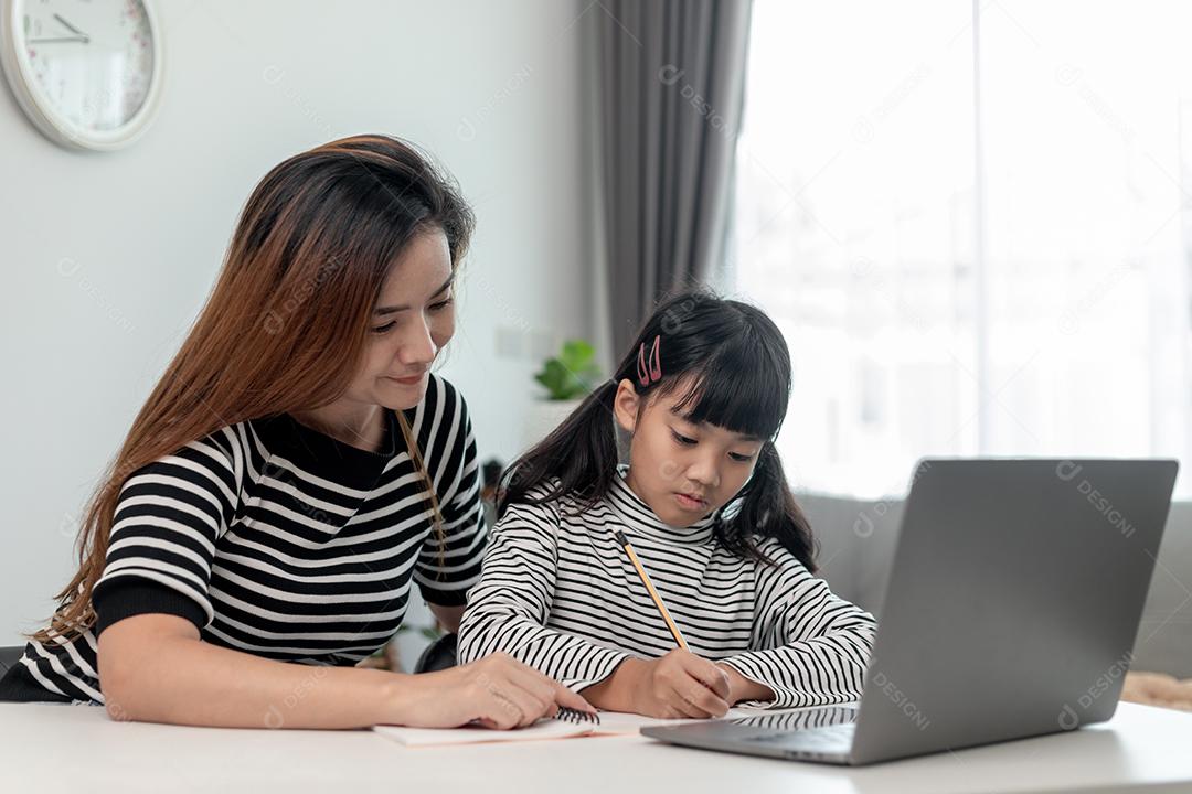 Garotinha asiática aprendendo aula online em casa com a mãe. A criança pré-escolar usa o laptop para fazer a lição de casa, o ensino em casa do professor da escola pela internet remota digital com o apoio da mãe.