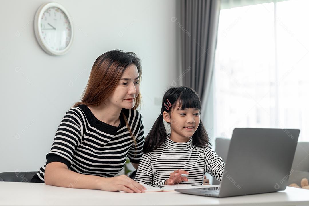 Garotinha asiática aprendendo aula online em casa com a mãe. A criança pré-escolar usa o laptop para fazer a lição de casa, o ensino em casa do professor da escola pela internet remota digital com o apoio da mãe.