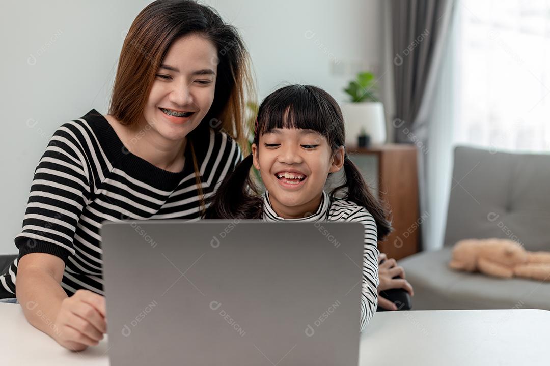 Garotinha asiática aprendendo aula online em casa com a mãe. A criança pré-escolar usa o laptop para fazer a lição de casa, o ensino em casa do professor da escola pela internet remota digital com o apoio da mãe.
