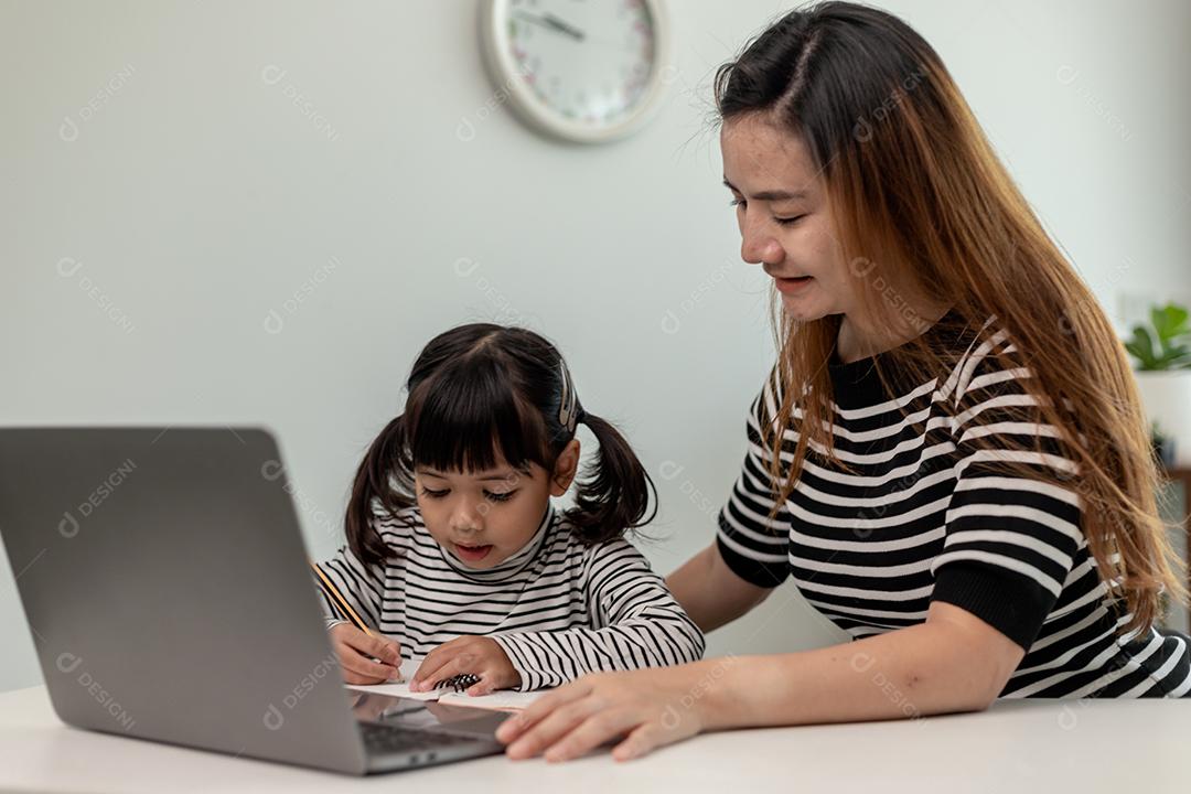 Garotinha asiática aprendendo aula online em casa com a mãe. A criança pré-escolar usa o laptop para fazer a lição de casa, o ensino em casa do professor da escola pela internet remota digital com o apoio da mãe.