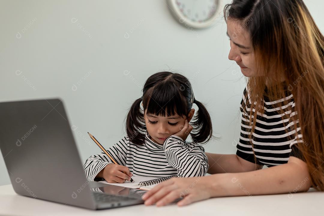 Garotinha asiática aprendendo aula online em casa com a mãe. A criança pré-escolar usa o laptop para fazer a lição de casa, o ensino em casa do professor da escola pela internet remota digital com o apoio da mãe.