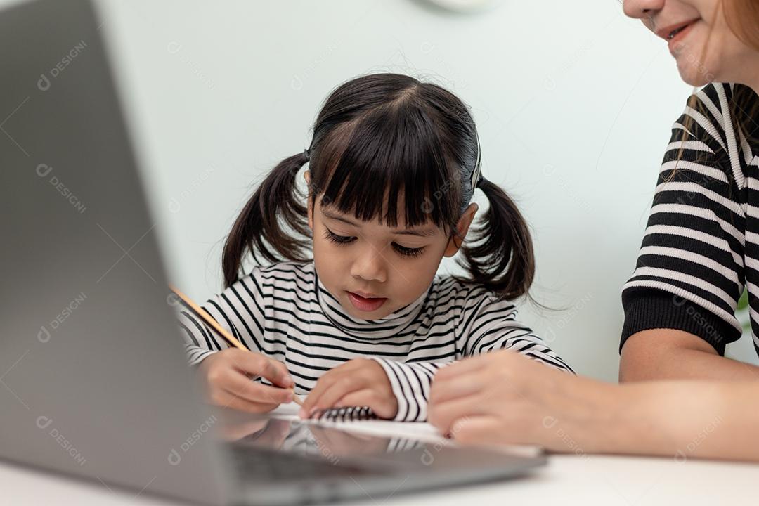 Garotinha asiática aprendendo aula online em casa com a mãe. A criança pré-escolar usa o laptop para fazer a lição de casa, o ensino em casa do professor da escola pela internet remota digital com o apoio da mãe.