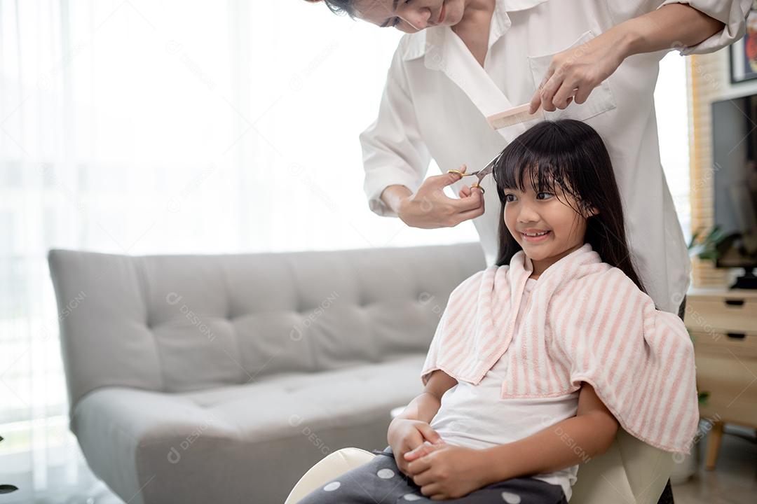 Mãe asiática cortando cabelo para a filha na sala de estar em casa enquanto fica em casa segura do Covid-19 Coronavirus durante o bloqueio. Conceito de auto-quarentena e distanciamento social.