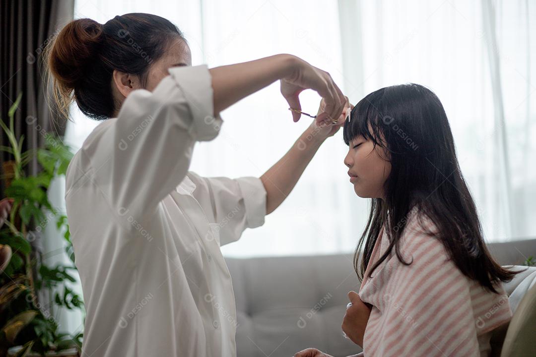 Mãe asiática cortando cabelo para a filha na sala de estar em casa enquanto fica em casa segura do Covid-19 Coronavirus durante o bloqueio. Conceito de auto-quarentena e distanciamento social.