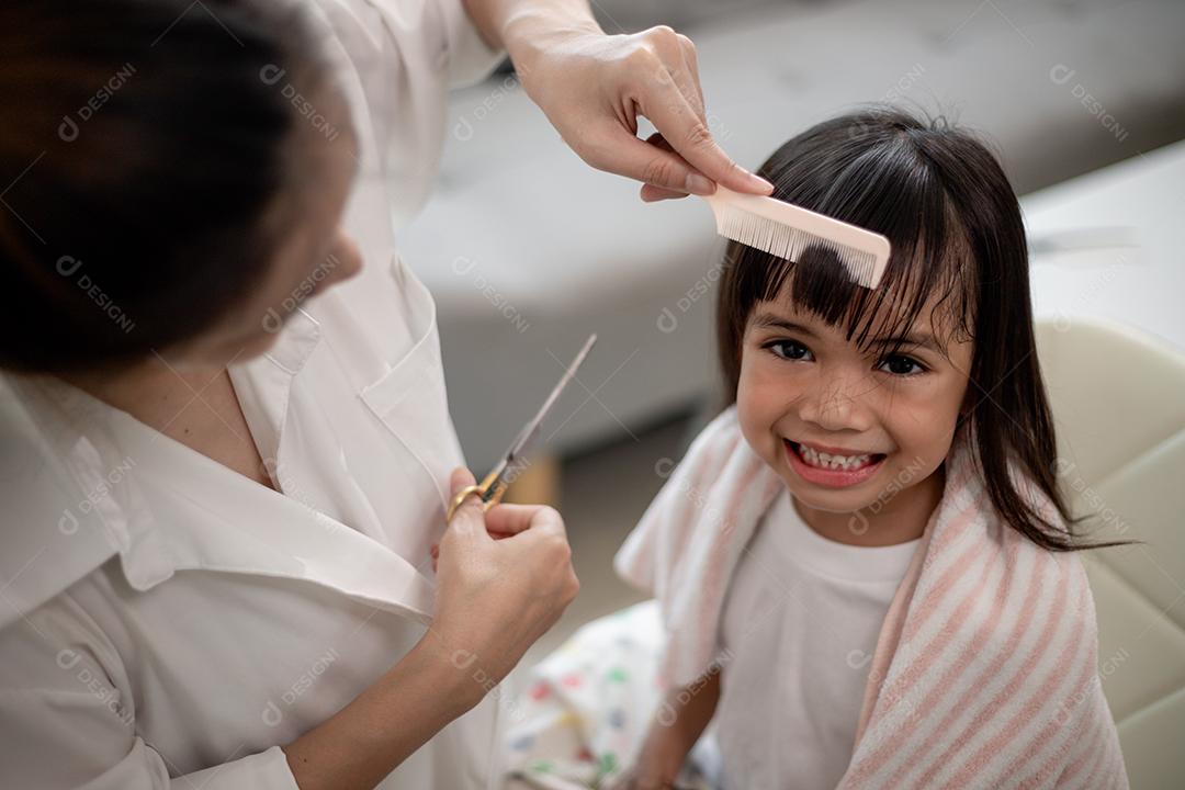 Mãe asiática cortando cabelo para a filha na sala de estar em casa enquanto fica em casa segura do Covid-19 Coronavirus durante o bloqueio. Conceito de auto-quarentena e distanciamento social.