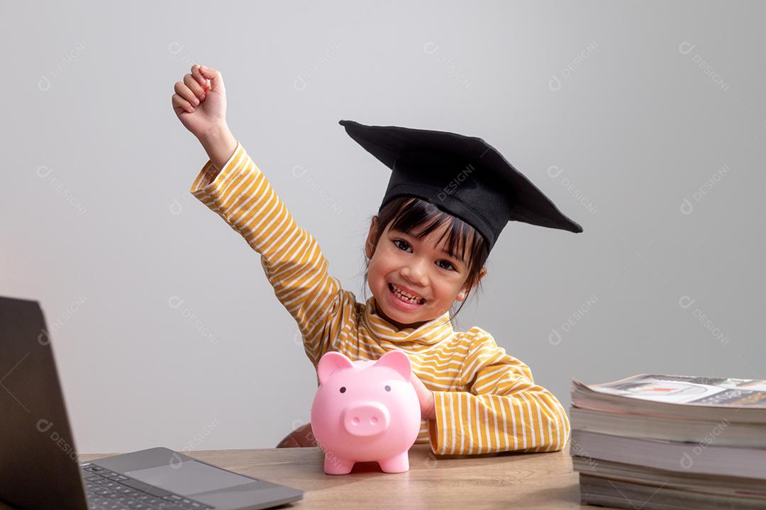 Menina asiática usando um chapéu de formatura com um cofrinho rosa, economizando dinheiro, investindo no futuro,