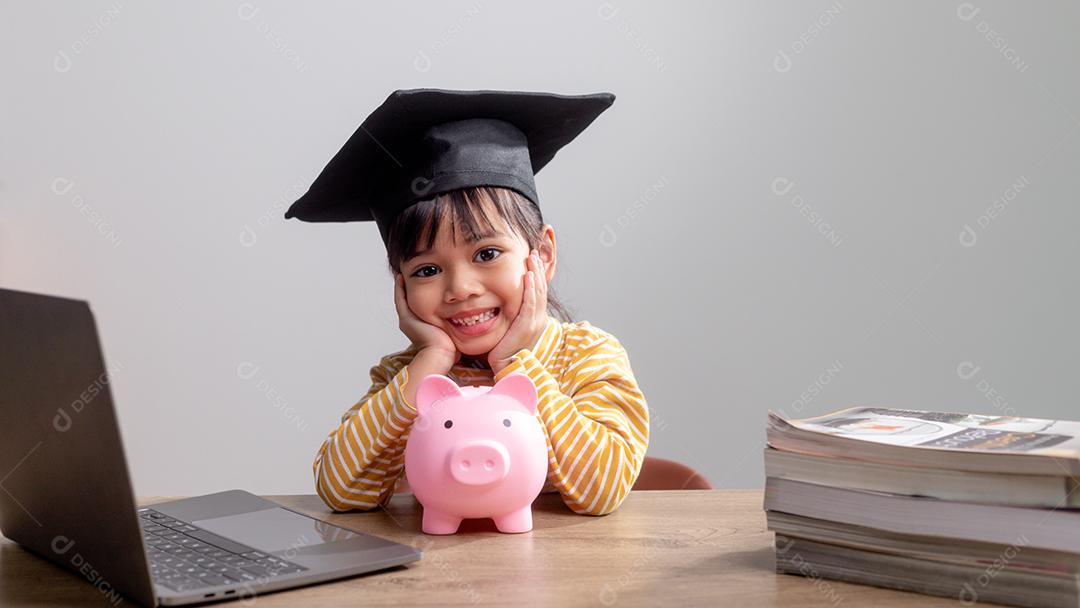 Menina asiática usando um chapéu de formatura com um cofrinho rosa, economizando dinheiro, investindo no futuro,
