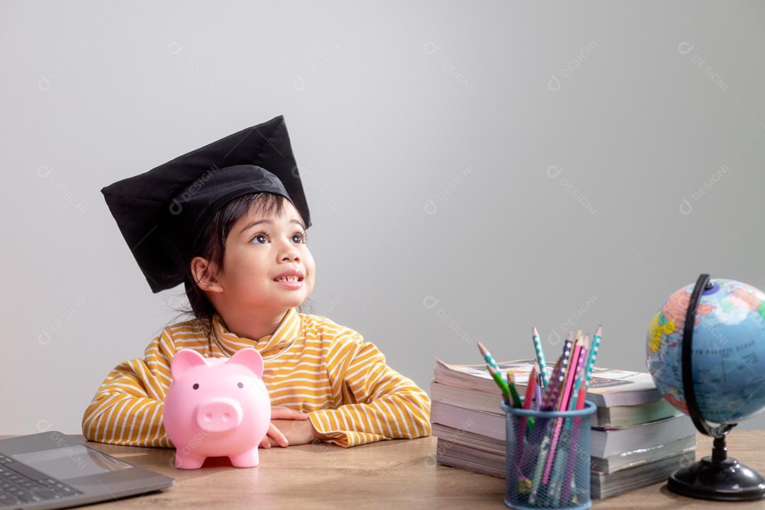Menina asiática usando um chapéu de formatura com um cofrinho rosa, economizando dinheiro, investindo no futuro,