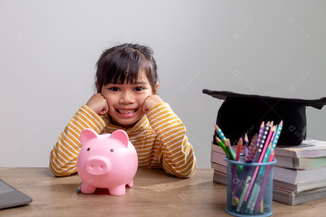 Menina asiática usando um chapéu de formatura com um cofrinho rosa, economizando dinheiro, investindo no futuro,