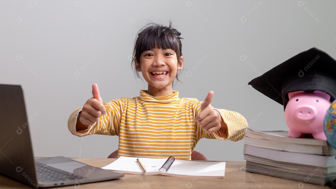 Menina asiática usando um chapéu de formatura com um cofrinho rosa, economizando dinheiro, investindo no futuro,