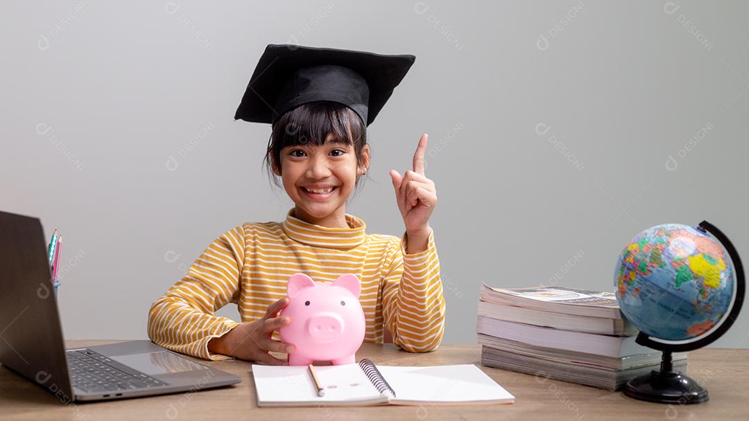 Menina asiática usando um chapéu de formatura com um cofrinho rosa, economizando dinheiro, investindo no futuro,