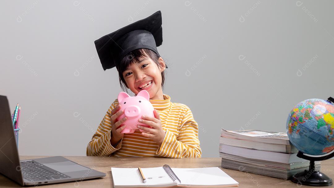 Menina asiática usando um chapéu de formatura com um cofrinho rosa, economizando dinheiro, investindo no futuro,