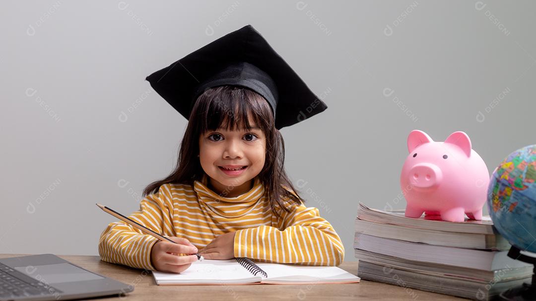 Menina asiática usando um chapéu de formatura com um cofrinho rosa, economizando dinheiro, investindo no futuro,