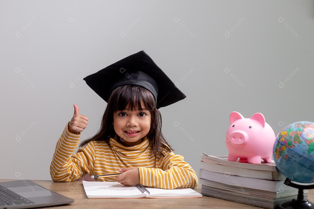 Menina asiática usando um chapéu de formatura com um cofrinho rosa, economizando dinheiro, investindo no futuro,