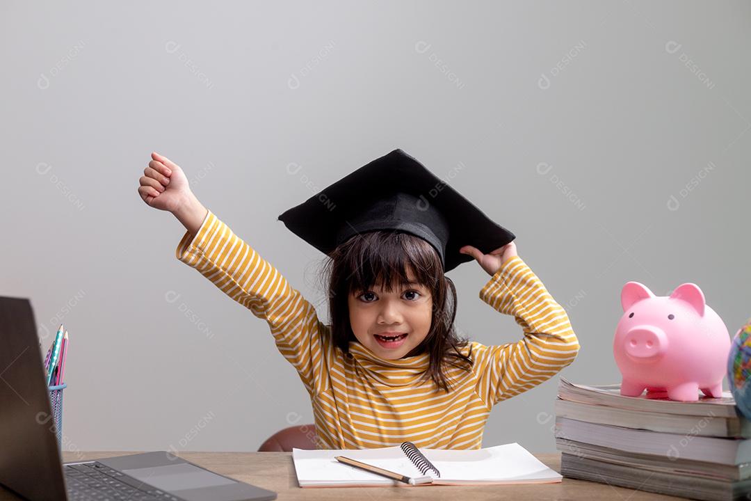 Menina asiática usando um chapéu de formatura com um cofrinho rosa, economizando dinheiro, investindo no futuro,