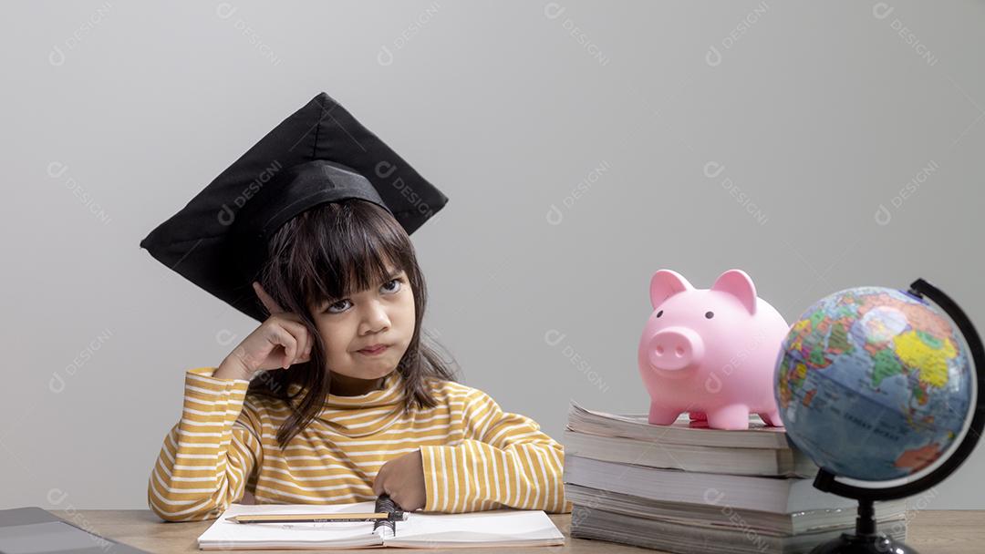 Menina asiática usando um chapéu de formatura com um cofrinho rosa, economizando dinheiro, investindo no futuro,