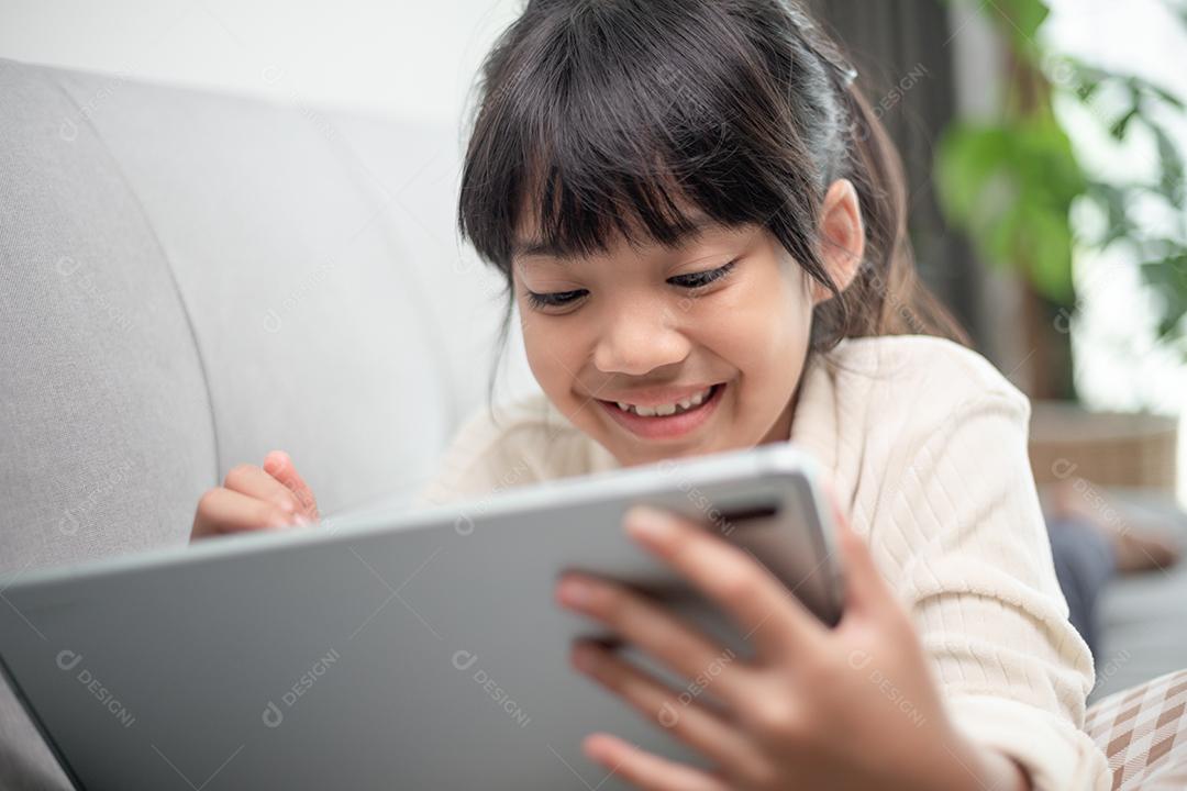 Menina usando tablet jogando na internet, criança sentada no sofá assistindo ou conversando com um amigo online, criança relaxando na sala de manhã, crianças com conceito de nova tecnologia