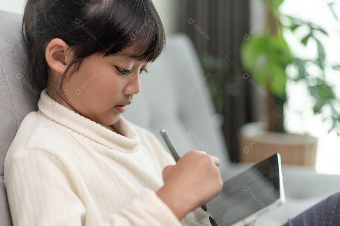 Menina usando tablet jogando na internet, criança sentada no sofá assistindo ou conversando com um amigo online, criança relaxando na sala de manhã, crianças com conceito de nova tecnologia