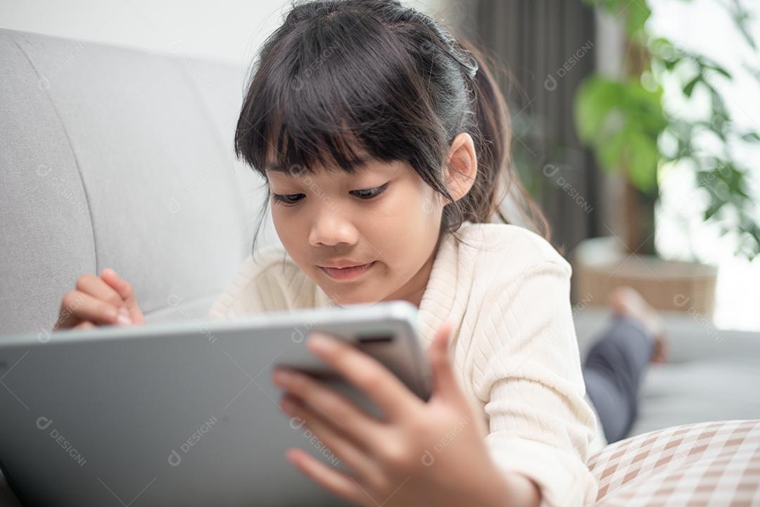 Menina usando tablet jogando na internet, criança sentada no sofá assistindo ou conversando com um amigo online, criança relaxando na sala de manhã, crianças com conceito de nova tecnologia