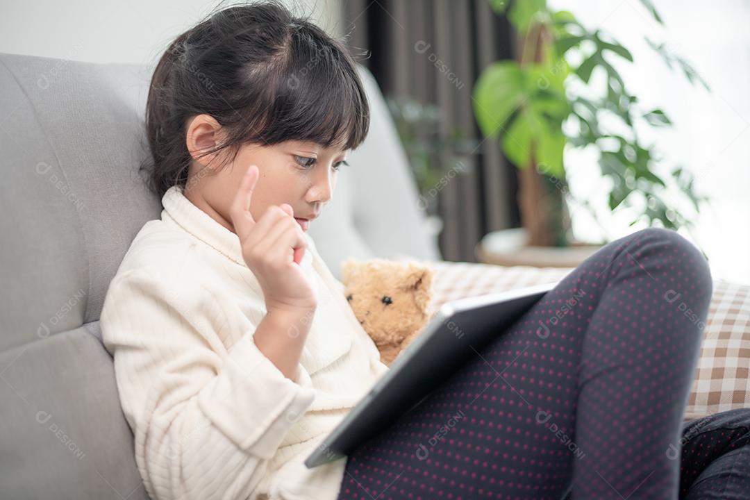 Menina usando tablet jogando na internet, criança sentada no sofá assistindo ou conversando com um amigo online, criança relaxando na sala de manhã, crianças com conceito de nova tecnologia