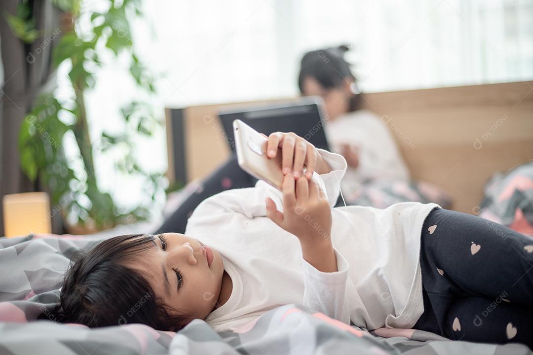 Garotinha bonitinha assistindo vídeo no smartphone com carinha sozinha na cama, criança usando celular com carinha feliz em casa. Fique em casa em quarentena coronavírus COVID-19 prevenção pandêmica.