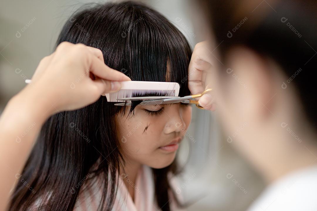 Mãe asiática cortando cabelo para a filha na sala de estar em casa enquanto fica em casa segura do Covid-19 Coronavirus durante o bloqueio. Conceito de auto-quarentena e distanciamento social.