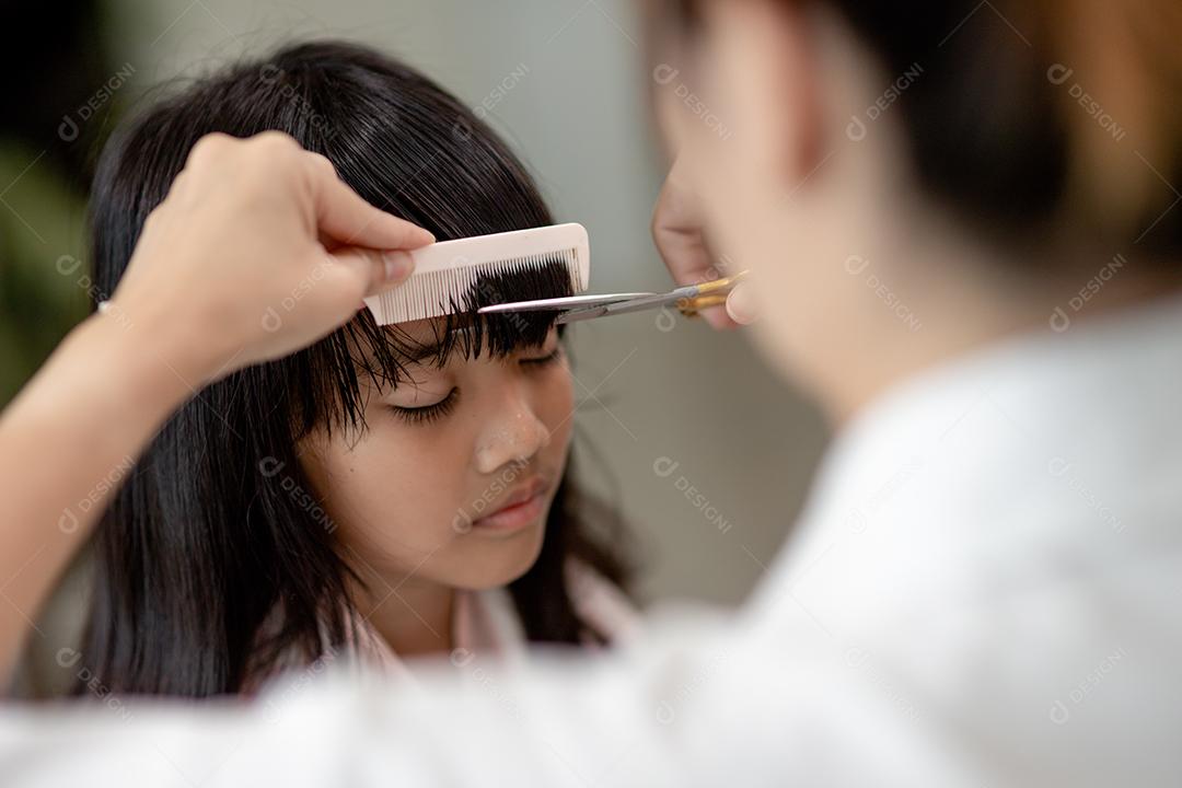 Mãe asiática cortando cabelo para sua filha na sala de estar em casa
