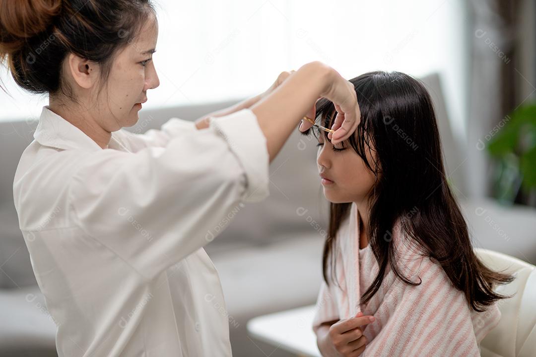 Mãe asiática cortando cabelo para a filha na sala de estar em casa enquanto fica em casa segura do Covid-19 Coronavirus durante o bloqueio. Conceito de auto-quarentena e distanciamento social.