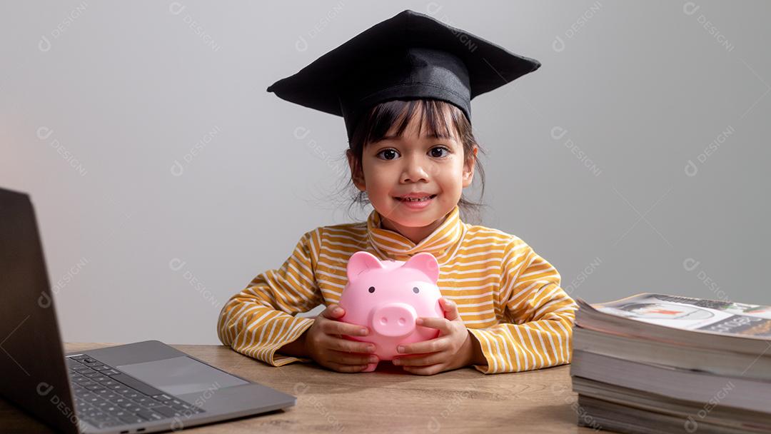 Menina asiática usando um chapéu de formatura com um cofrinho rosa, economizando dinheiro, investindo no futuro,