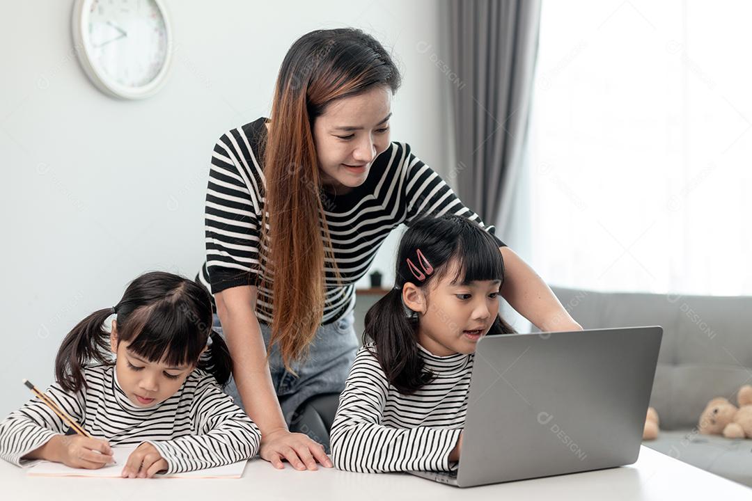 Garotinha asiática aprendendo aula online em casa com a mãe. A criança pré-escolar usa o laptop para fazer a lição de casa, o ensino em casa do professor da escola pela internet remota digital com o apoio da mãe.