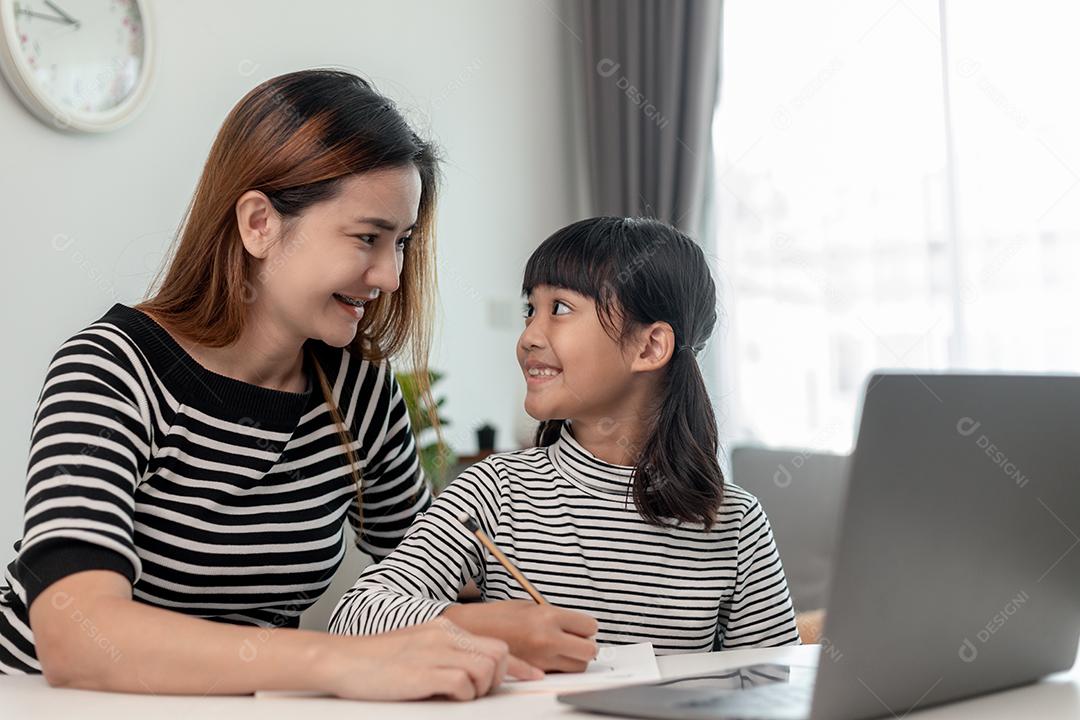 Garotinha asiática aprendendo aula online em casa com a mãe. A criança pré-escolar usa o laptop para fazer a lição de casa, o ensino em casa do professor da escola pela internet remota digital com o apoio da mãe.