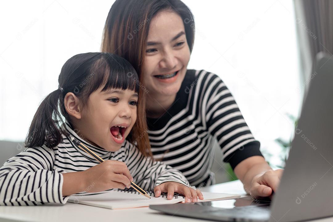 Garotinha asiática aprendendo aula online em casa com a mãe. A criança pré-escolar usa o laptop para fazer a lição de casa, o ensino em casa do professor da escola pela internet remota digital com o apoio da mãe.