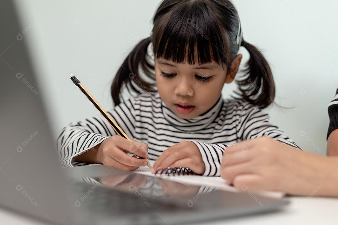 Garotinha asiática aprendendo aula online em casa com a mãe. A criança pré-escolar usa o laptop para fazer a lição de casa, o ensino em casa do professor da escola pela internet remota digital com o apoio da mãe.